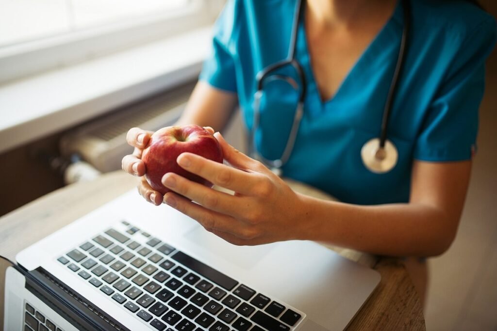 Soignante en tenue bleue, stéthoscope au cou, assise devant un ordinateur portable une pomme à la main