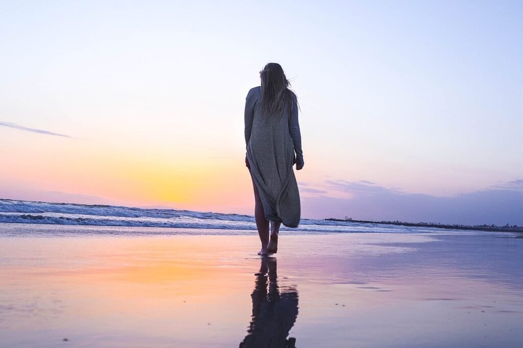 Femme vue de dos marchant pieds nus au bord de l'eau sur une plage au coucher du soleil