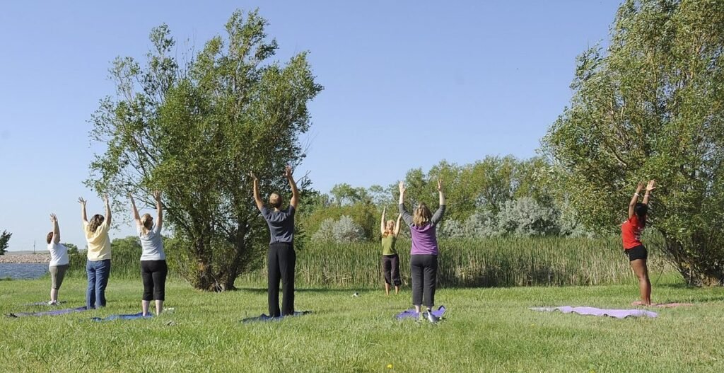 Groupe pratiquant le yoga bras levés sur une pelouse au bord de l'eau, sous un ciel bleu