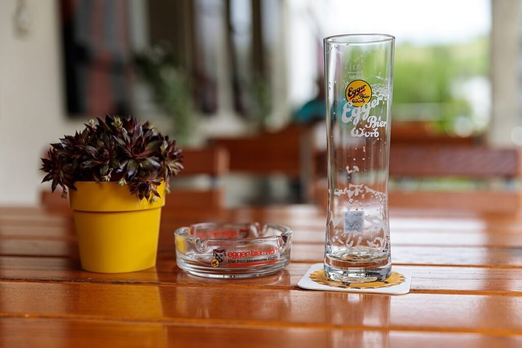 Cendrier en verre vide sur une table de terrasse en bois, à côté d'un pot de fleurs jaune
