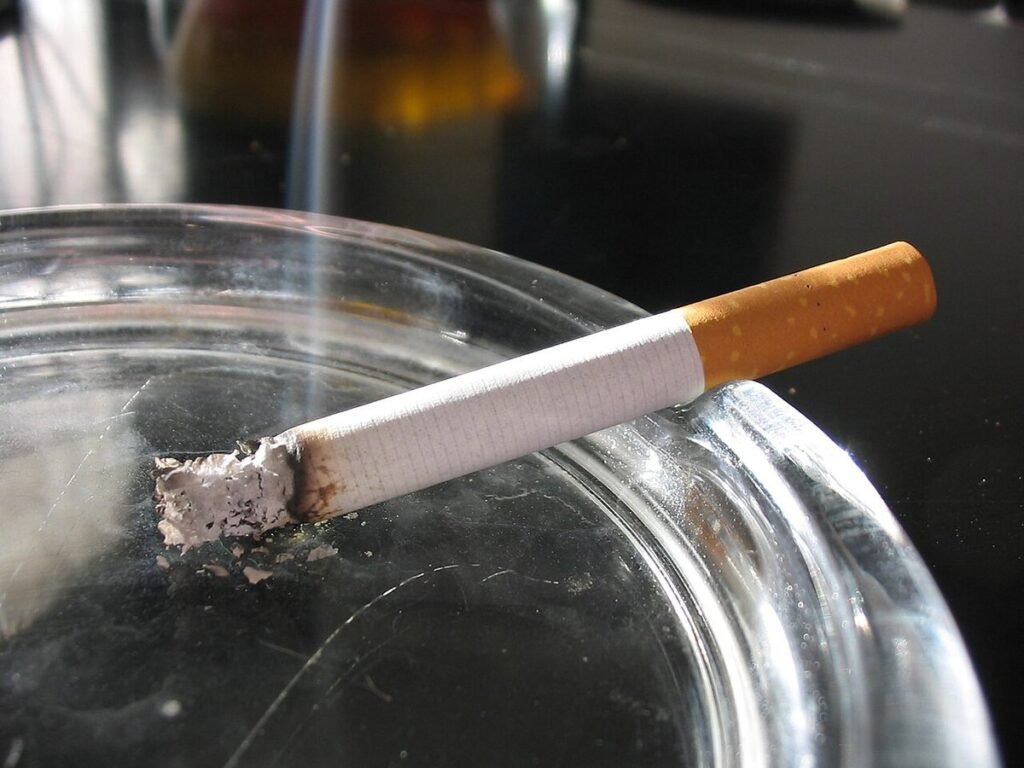 Cigarette allumée posée sur le bord d'un cendrier en verre, un filet de fumée s'élevant